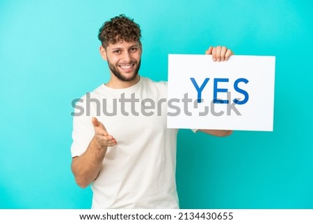 Young handsome caucasian man isolated on blue background holding a placard with text YES making a deal