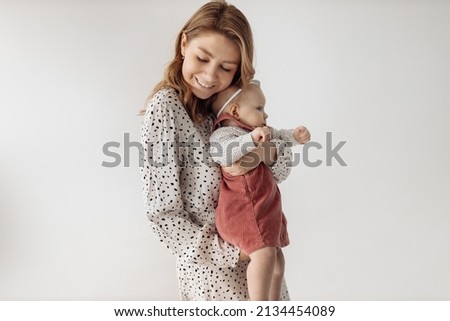 A beautiful young mother holds her adorable daughter in her arms, hugging her. A cute little girl looks at the camera, mom gently looks at her baby, smiles. Happy motherhood concept. 