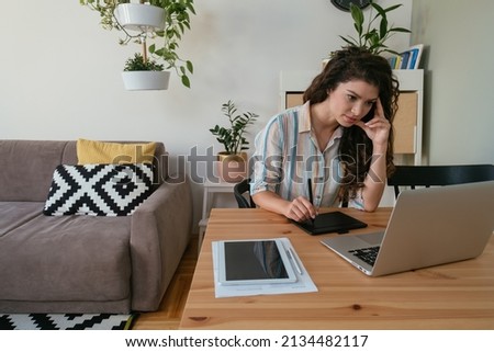 Tired Business Woman Working at Home Office on her Laptop Computer and Retouching Pad. 
Smiling female graphic designer drawing something on a graphic tablet while sitting at desk in the living room.