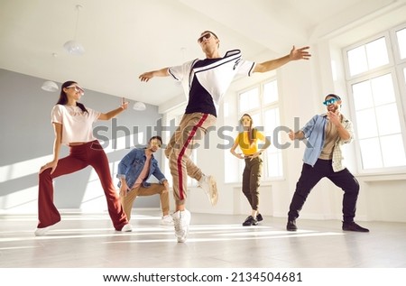 Expressive contemporary hip hop dance. Young male dancer performs hip hop movement on one leg during break dance lesson. Guy dances on surrounded by his dance friends in bright hall of dance studio. Royalty-Free Stock Photo #2134504681