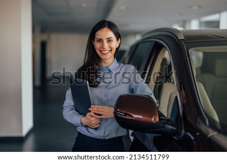 Picture of lovely brunette woman, leaning onto the car while carrying her laptop.