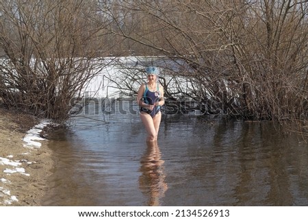 An adult woman with magnolia flower in blue kokoshnik walks through cold water in spring against the background of melting ice floes and bushes on sunny day.