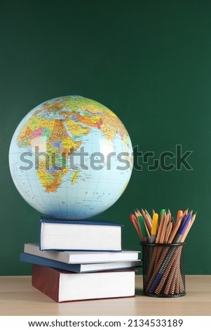 Globe, school supplies and books on wooden table near green chalkboard. Geography lesson