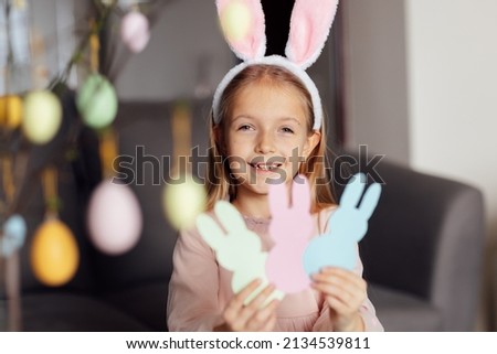 happy caucasian toddler girl eight years old at home in living room making garland with paper bunny. Stay home during Coronavirus covid-19 pandemic