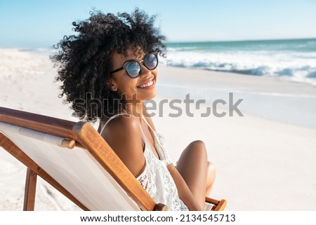 Portrait of happy young black woman relaxing on wooden deck chair at tropical beach while looking at camera wearing spectacles. Smiling african american girl with fashion sunglasses enjoying vacation. Royalty-Free Stock Photo #2134545713