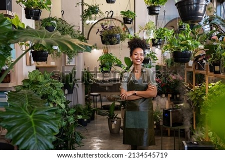 Portrait of african american woman with crossed arms wearing apron in botanical store. Happy small business owner working at flower shop standing surrounded by plants while looking at camera. Royalty-Free Stock Photo #2134545719