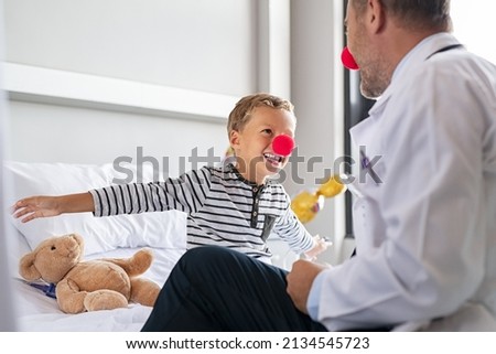 Smiling little boy wearing red nose and flying with outstretched arms like superhero in hospital bed with doctor. Kid joking during medical examination with physician, clown therapy concept.  Royalty-Free Stock Photo #2134545723