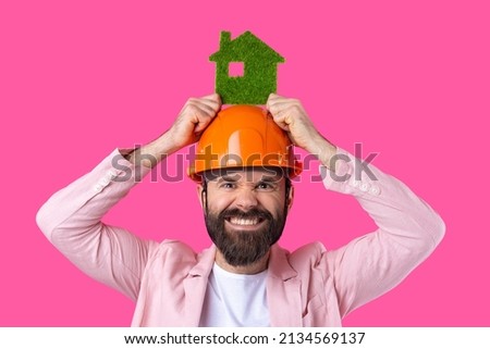 Portrait of young construction engineer wear orange hard hat, in a pink jacket standing on red studio background. A man holds a green eco house.