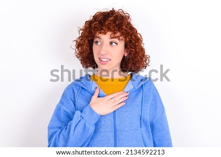 Joyful young redhead girl wearing blue jacket over white background  expresses positive emotions recalls something funny keeps hand on chest and giggles happily.