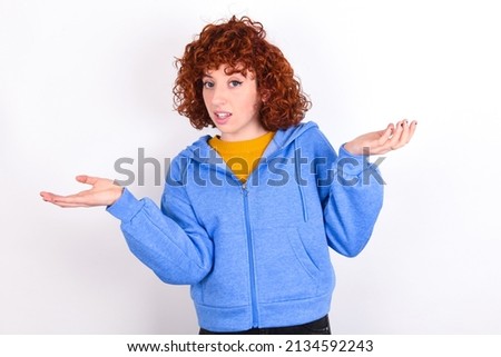 So what? Portrait of arrogant young redhead girl wearing blue jacket over white background  shrugging hands sideways smiling gasping indifferent, telling something obvious. Royalty-Free Stock Photo #2134592243