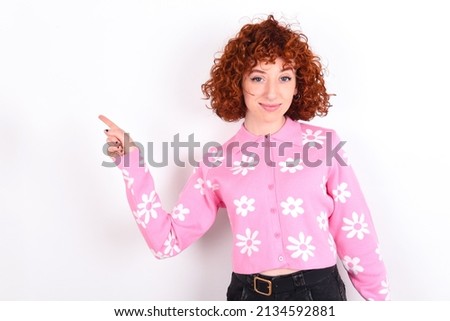 Positive young redhead girl wearing pink floral t-shirt over white background with satisfied expression indicates at upper right corner shows good offer suggests to click on link Royalty-Free Stock Photo #2134592881