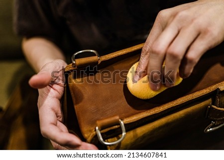 Professional leather care service concept. Closeup hand with yellow soft sponge cleaning stains on the surface of the vintage brown bag, depth of field. Polish waxing protection from dust and dirt. Royalty-Free Stock Photo #2134607841
