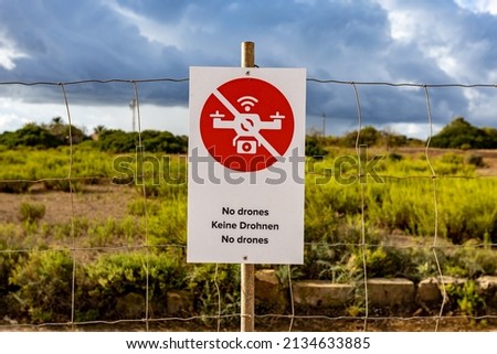 a white sign with a red prohibition sign saying "no drones". Sign on a fence indicating a no-fly zone. overcast gray sky. 