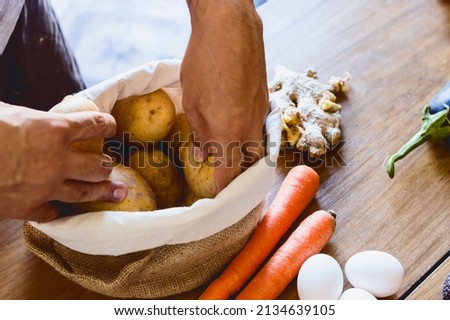 closeup of a man standing in the kitchen of his house selecting potatoes that are inside a sack along with other vegetables on the table