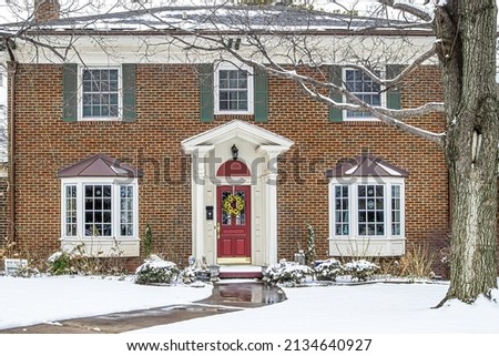 Red brick house in snow with bay windows and columns and green shutters and a sunflower wreath