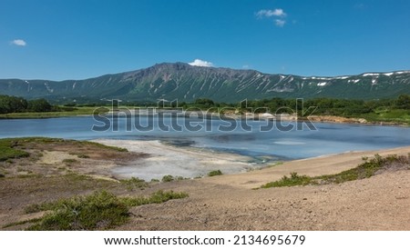 Turquoise thermal lake in the caldera of an extinct volcano. There are sulfur deposits on the soil. Green vegetation on the shore. A picturesque mountain range against the blue sky. Kamchatka. Uzon