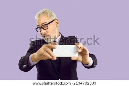 Senior businessman making funny face and taking selfie on mobile phone. Studio shot of handsome white haired mature man in glasses holding smartphone, making silly grimace and taking photo of himself