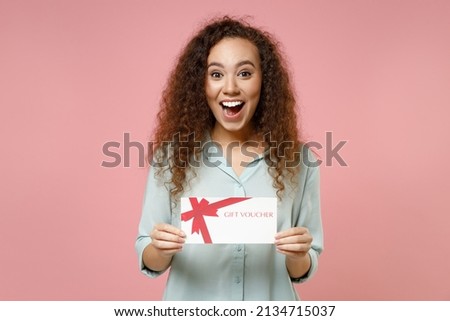 Young black african american surprised shocked cute rich curly woman 20s wearing blue shirt holding gift voucher flyer mock up looking camera isolated on pastel pink color background studio portrait