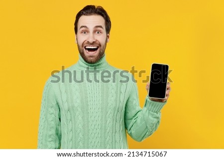 Young smiling happy cheerful fun cool man 20s wear mint knitted sweater hold in hand use mobile cell phone with blank screen workspace area isolated on plain yellow color background studio portrait.
