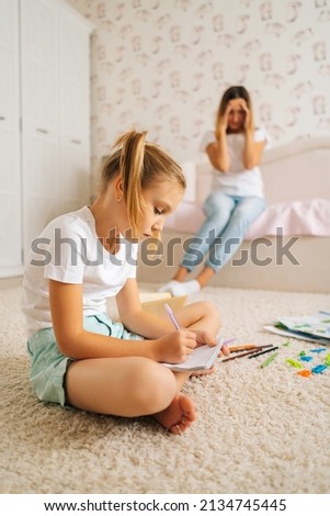 Vertical shot of primary girl child writing homework in copybook sitting on floor with laptop for distance education online at home, on blurred background of shocked young mother sitting on sofa.