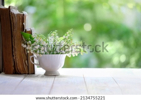 Lily of the valley flowers in cup and books on white table, green natural background. symbol of spring season. beautiful romantic floral composition. copy space Royalty-Free Stock Photo #2134755221