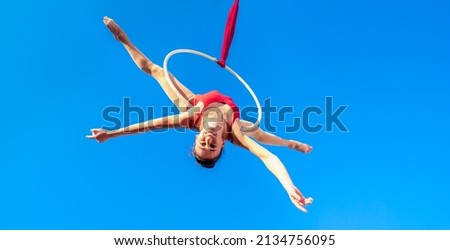 acrobat athletic, young graceful gymnast performing aerial exercise in the air ring outdoors on sky background. flexible woman in red suit performs circus artist dancing on hoop upside down. low angle