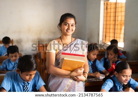Smiling Teacher with books in hand standing at classroom in between students at classroom - concept of professional occupation, woman empowerment and education Royalty-Free Stock Photo #2134787565