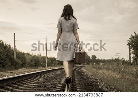 A rear view of a young woman in a dress and with a suitcase walking away along the rails of a railway road. The concept of departure, emigration, departure from the country, refugee status,deportation Royalty-Free Stock Photo #2134793867