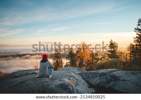 Hiker sits atop Vuokatinvaara in Sotkamo in the Kainuu region, Finland at sunrise and above the clouds. A brunette woman with a red knitted hat on top of the mountain. Royalty-Free Stock Photo #2134810025