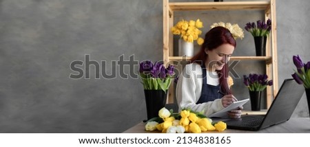 Portrait of a florist in an apron, working in her own flower shop, using a laptop organizing logistics and delivery, taking orders online, taking notes with a pen in a notebook. Banner. copy space.
