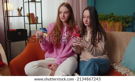 Cheerful girls friends siblings using credit bank card and smartphone while transferring money, purchases online shopping. Happy two female women couple family on sofa at home, spending time together
