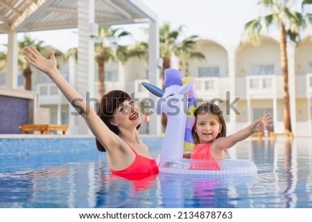 Young beautiful woman and her little daughter swimming in the pool of exotic luxury resort. A girl in a life-saving inflatable unicorn enjoying relaxing in the water with her mother.