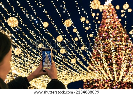 A woman taking photo of tall urban festive Christmas tree on smartphone, bright blurred background with bokeh lights.
