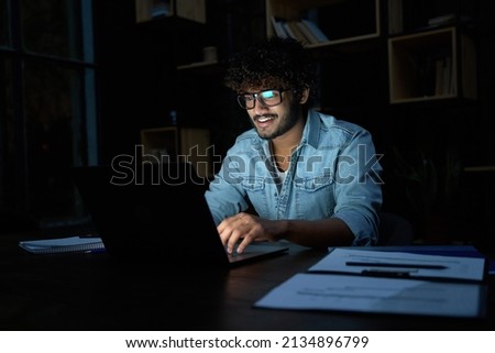 Smiling young curly indian latin ethnic business man or student wearing glasses remote working overtime, learning online late at night at home or in dark office using laptop computer at workplace. Royalty-Free Stock Photo #2134896799