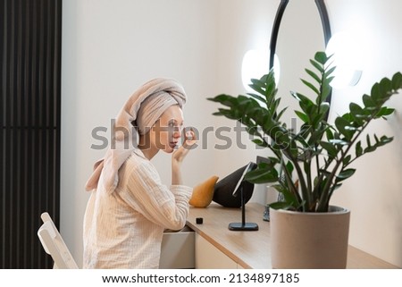 profile photo of young caucasian woman sitting at her makeup table, looking at big mirror, wet hair wrapped in towel, after shower, taking care of herself, applying moisturising cream, green plant