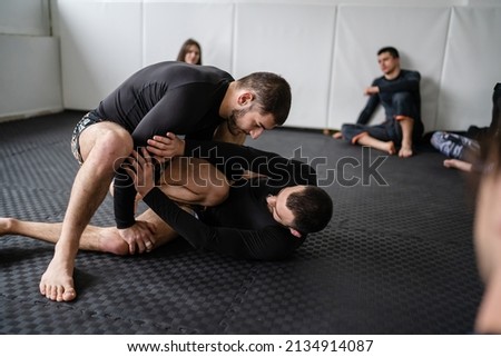 two men demonstrate bjj brazilian jiu jitsu grappling or luta livre technique on the ground at training at the academy in front of group of students leg attack Royalty-Free Stock Photo #2134914087