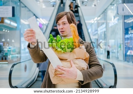 Man in a coat holds a paper check and a bag with a baguette and lettuce in a shopping mall and is shocked by the high prices of groceries. Royalty-Free Stock Photo #2134928999