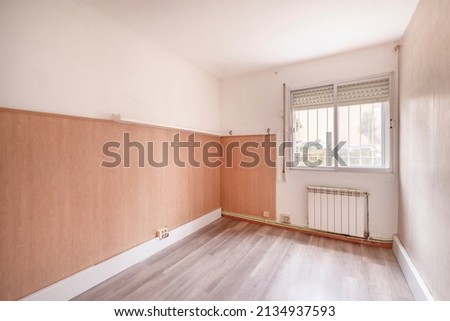 Empty living room with window and white aluminum radiator below, gray painted walls, white rogapie and wooden floorboards