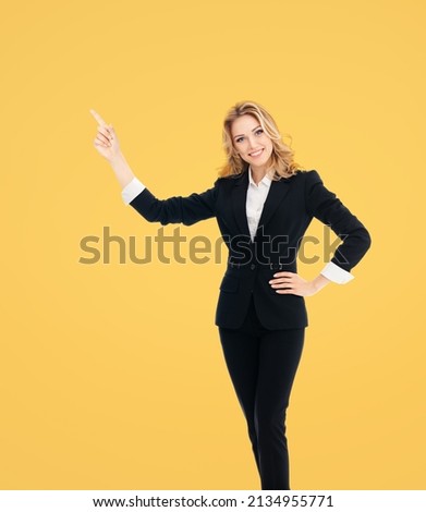 Full body of smiling executive woman in black confident suit, pointing showing advertising copy space copyspace. Business ad concept. Yellow background. Young cheerful businesswoman. Advertiser lady.