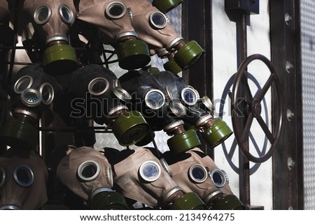 A full frame dark image of rows of gas masks with bright green filters on them and blue glass eyepieces. Bunker with round valve is on the side of the frame. Dark scary war picture