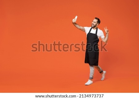 Full length young man barista bartender barman employee in black apron white t-shirt work coffee shop do selfie shot on mobile phone show v-sign isolated on orange background. Small business startup