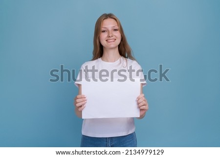 Young woman holding white paper on blue background. Caucasian woman with white blank template sheet with empty space in white t-shirt and blue jeans shows advertisement, mockup