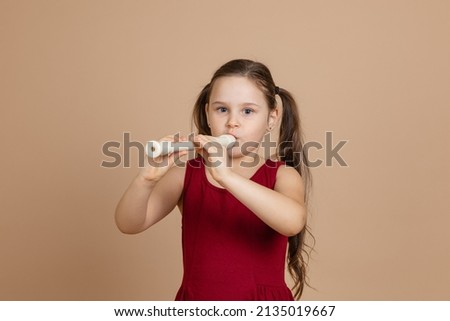 Girl in red dress thoughtfully play melody on flute, blowing air into duct, beige background. Learn to play woodwind musical instrument. Flute and children is concept of music education development. Royalty-Free Stock Photo #2135019667