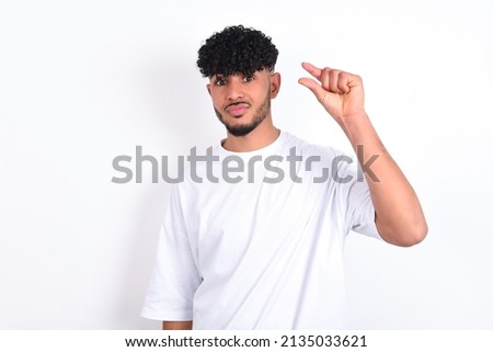 Upset young arab man wearing white T-shirt over white background shapes little gesture with hand demonstrates something very tiny small size. Not very much