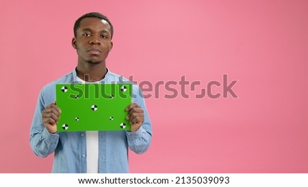 Serious African-American holding Banner with Green Screen Tracks Points for Copy Space. Empty Green Screen Board. Does Not Smile Looks at Camera With Space for Text or Advertising on Pink Background.
