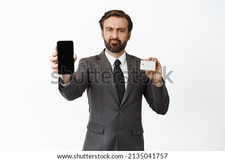 Disappointed corporate man showing smth upsetting on mobile phone screen and holding credit card, furrow eyebrows and grimacing displeased, white background