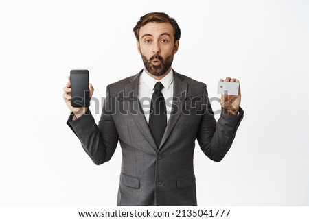 Businessman looking amazed, showing telephone screen and credit card, advertising app, shopping application, standing over white background