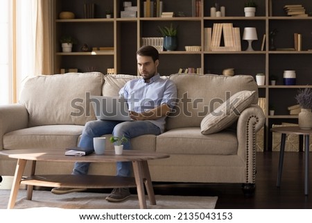 Serious guy freelance worker remote student sit on comfy couch at home do job learn online on distance hold laptop on knees. Busy young male write email type message prepare electronic document on pc
