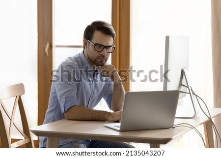 Serious young man in glasses work from home office on two computers testing software practicing currency trading. Thoughtful millennial businessman processing data speculating on stock market using pc Royalty-Free Stock Photo #2135043207