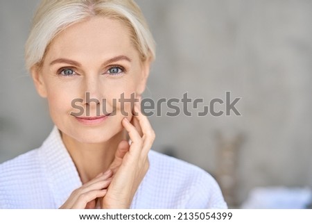 Closeup of happy smiling beautiful senior adult woman of 50s wearing bathrobe at spa hotel looking at camera touching face. Antiage spa procedures advertising. Skin care products concept. Copy space. Royalty-Free Stock Photo #2135054399